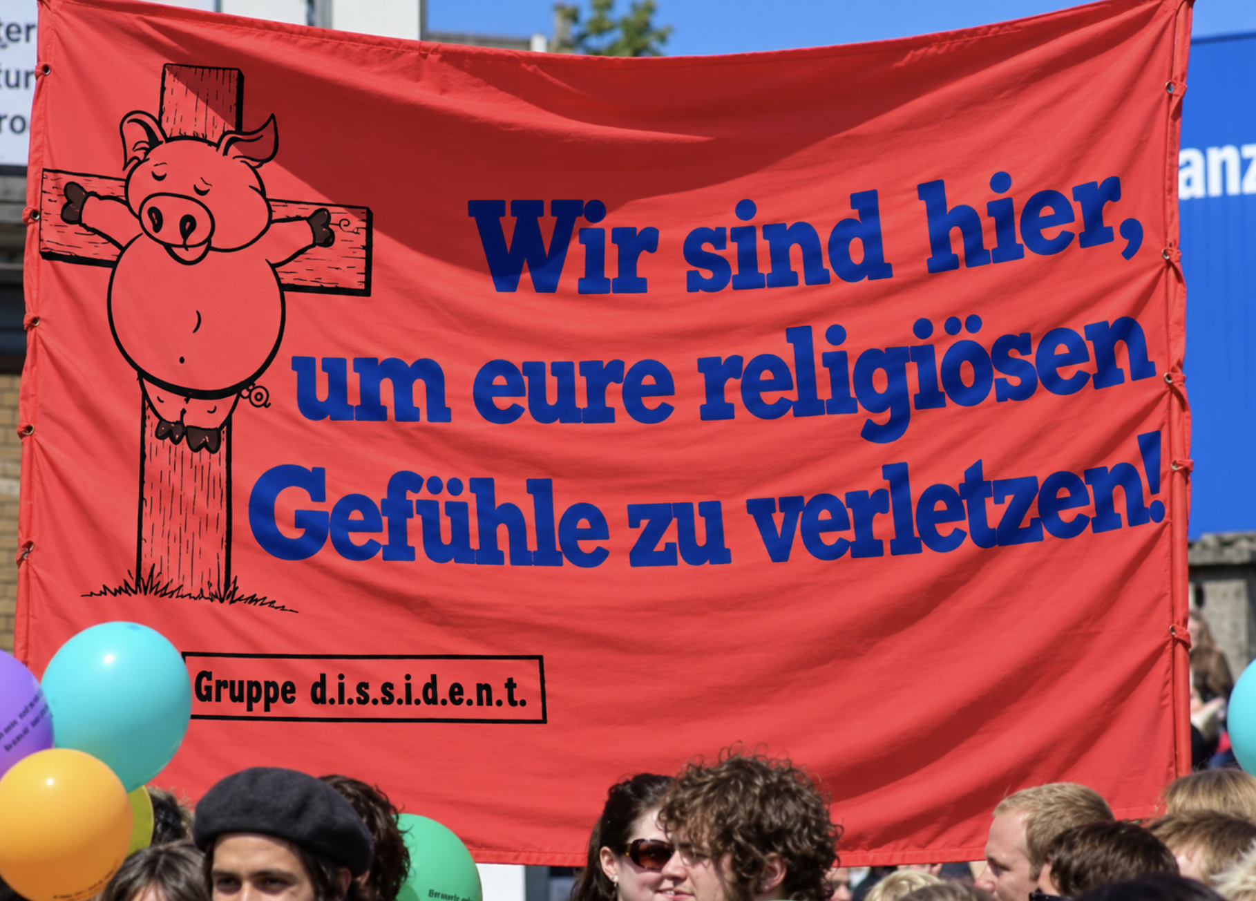 Rotes Banner mit einem Schwein und der Aufschrift „Wir sind hier, um eure religiösen Gefühle zu verletzen!“ bei einer Demonstration.